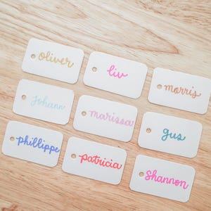 May include: Nine white rectangular tags with rounded corners, each featuring a different name in colorful cursive script. Names include Oliver, Liv, Morris, Johann, Marissa, Gus, Phillippe, Patricia, and Shannon. Each tag has a small hole.