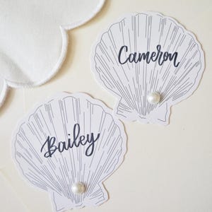 May include: Two white paper shell-shaped place cards featuring black line art details. Each card displays a name in black script, with 'Cameron' on one and 'Bailey' on the other. A single small white pearl is attached to the bottom of each shell.