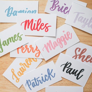 May include: A collection of white rectangular cards with names written in various colors of cursive script. Names include Damian, Miles, Maggie, Patrick, and Paul. The cards are scattered on a light brown wooden surface.