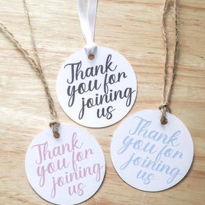 May include: Three white, round gift tags with the text "Thank you for joining us" in black, pink, and blue script. Each tag has a hole at the top with a ribbon or twine for hanging. The tags are on a wooden surface.