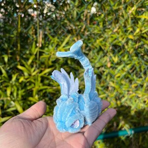 May include: A light blue pipe cleaner creature with a long tail and wings. The creature has a sparkly blue eye and is sitting on a hand.