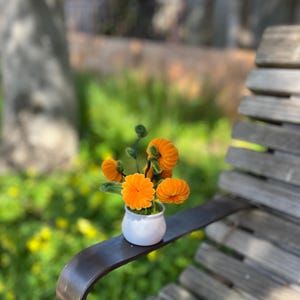 Pode incluir: Um pequeno vaso de cerâmica branca com um buquê de flores de feltro laranja. O vaso está sentado em um banco de madeira.
