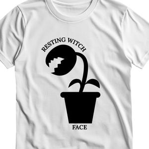 Op de afbeelding: Een wit T-shirt met korte mouwen en een zwarte afbeelding van een potplant met een mond met gekartelde randen. De tekst "RESTING WITCH" is in een zwarte boog boven de plant gedrukt en het woord "FACE" staat in het zwart onder de pot.