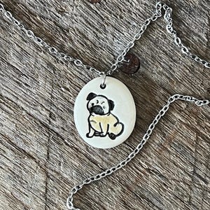 May include: A silver-toned necklace featuring a pendant with a hand-drawn pug illustration. The pendant is an oval shape with a cream-colored background. The pug is depicted in black and tan, with a detailed outline.