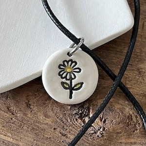 May include: A round white ceramic pendant featuring a hand-drawn black outline of a daisy with a yellow center and green leaves. The pendant is attached to a thin black cord necklace by a silver-toned metal jump ring, creating a simple accessory.