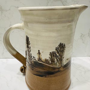 Puede incluir: Jarra de cerámica hecha a mano con un diseño de paisaje. La jarra presenta una parte superior de color crema, una base marrón y un asa. El diseño del paisaje incluye árboles y colinas en tonos marrones y negros. La jarra mide aproximadamente 25 cm de altura.