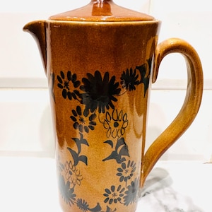 May include: Brown ceramic coffee pot with a lid and handle. The pot is decorated with black floral designs. The pot has a spout for pouring. The coffee pot is on a white surface.