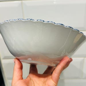 May include: A white ceramic bowl with a smooth, glossy finish and a scalloped rim. The edge of the bowl features a detailed, intricate blue and white pattern that contrasts with the plain white interior and exterior of the basin.
