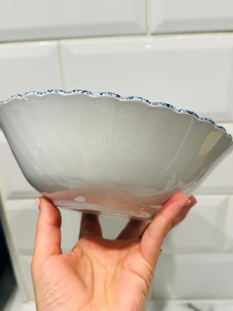 May include: A white ceramic bowl with a smooth, glossy finish and a scalloped rim. The edge of the bowl features a detailed, intricate blue and white pattern that contrasts with the plain white interior and exterior of the basin.