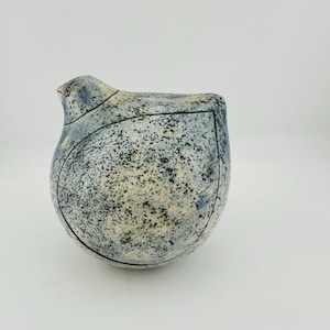Può includere: Una statuetta di uccello in ceramica arrotondata con una smaltatura blu e bianca maculata. La superficie è coperta da fini macchie blu scuro e nere, con distinte linee incise scure che delineano la forma dell'ala e della testa sul corpo.