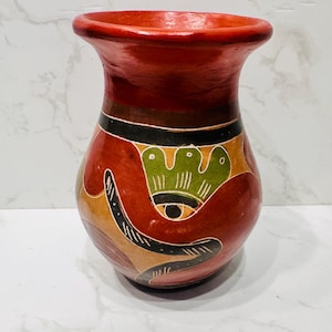 Puede incluir: Un jarrón de cerámica roja hecho a mano, con una abertura ancha y un cuerpo bulboso. El jarrón presenta un diseño pintado con detalles marrones, verdes y negros. El diseño incluye un ojo estilizado y elementos florales. El jarrón es decorativo.