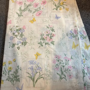 May include: Cream-coloured fabric with a floral and butterfly design. The pattern includes framed illustrations of pink, yellow, and purple flowers, interspersed with yellow and blue butterflies. The fabric is folded, revealing multiple layers.
