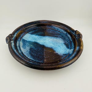 May include: A round ceramic plate featuring a dark brown base glaze with a prominent, textured light blue glaze accent across the centre. The plate has a raised rim and two small, sculpted handles on opposite sides with a glossy finish.