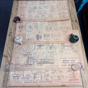Puede incluir: Una mesa de madera exhibe planos antiguos de un modelo de motor de tracción en funcionamiento, con el texto "Working Model Traction Engine". Una cinta métrica, dos tazas de té pequeñas y dos vasos transparentes también están sobre la mesa.