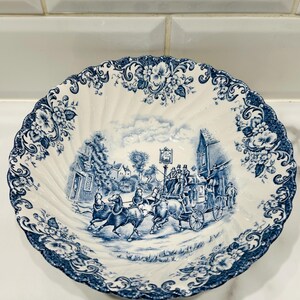 May include: A white ceramic bowl with a scalloped edge and a blue transferware pattern. The center features a detailed scene of a horse-drawn carriage in a village, while the rim is decorated with a dense floral motif in matching blue ink.