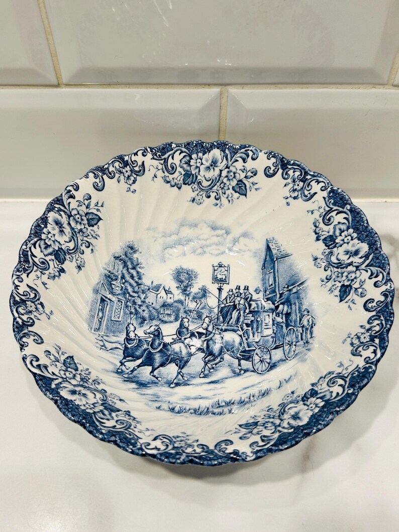 May include: A white ceramic bowl with a scalloped edge and a blue transferware pattern. The center features a detailed scene of a horse-drawn carriage in a village, while the rim is decorated with a dense floral motif in matching blue ink.