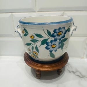 May include: A white ceramic plant pot with blue floral patterns and a blue rim, placed on a small wooden stand. The pot has two small handles and is decorated with blue and yellow flowers and green leaves.