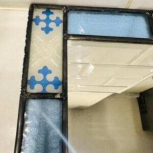 K&ouml;nnte beinhalten: Ein Eckabschnitt eines Spiegels, gerahmt in dunklem Metall mit gel&ouml;teten Verbindungen. Der Rahmen zeigt rechteckige Paneele aus strukturiertem hellblauem Glas und ein wei&szlig;es Glaspaneel, verziert mit zwei blauen Kreuzsymbolen und &Auml;tzmustern.