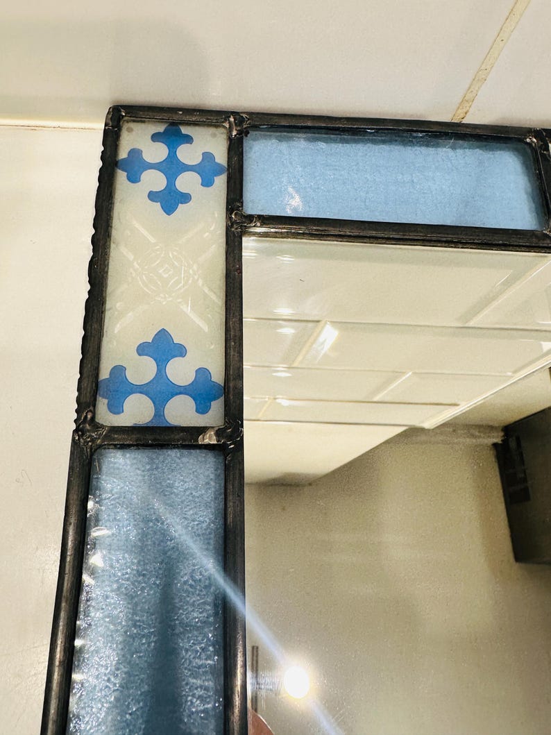 K&ouml;nnte beinhalten: Ein Eckabschnitt eines Spiegels, gerahmt in dunklem Metall mit gel&ouml;teten Verbindungen. Der Rahmen zeigt rechteckige Paneele aus strukturiertem hellblauem Glas und ein wei&szlig;es Glaspaneel, verziert mit zwei blauen Kreuzsymbolen und &Auml;tzmustern.