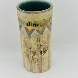 May include: A cylindrical ceramic vase with a teal interior. The exterior features a cream-coloured base with vertical tan stripes and black speckles. The upper section is decorated with a band of geometric triangles in yellow, blue, and brown.