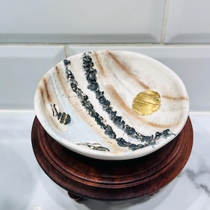 Può includere: Una ciotola ovale in ceramica con un motivo marmorizzato bianco, marrone e nero, impreziosita da un cerchio in foglia d'oro e una piccola illustrazione di un'ape. La ciotola è su un supporto in legno marrone scuro.