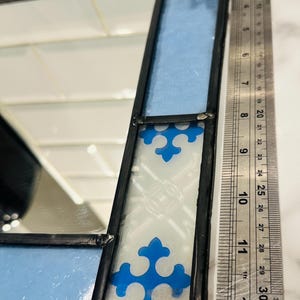 K&ouml;nnte beinhalten: Ein Buntglasspiegel mit hellblauen und wei&szlig;en Paneelen sowie blauen Kreuzmotiven, eingefasst in schwarzes Metall. Ein Edelstahl-Lineal liegt neben dem Spiegel und zeigt eine L&auml;nge von etwa 30 Zentimetern an.