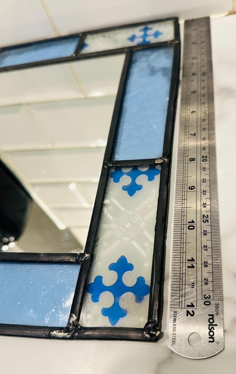 K&ouml;nnte beinhalten: Ein Buntglasspiegel mit hellblauen und wei&szlig;en Paneelen sowie blauen Kreuzmotiven, eingefasst in schwarzes Metall. Ein Edelstahl-Lineal liegt neben dem Spiegel und zeigt eine L&auml;nge von etwa 30 Zentimetern an.