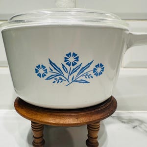 May include: A white saucepan with a glass lid and a handle. The saucepan features a blue floral design. It sits on a small, round, wooden stand with four legs. The background is a white tiled wall.