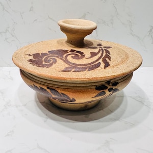 Può includere: Una ciotola in ceramica beige con coperchio. Il coperchio presenta un manico e un motivo floreale marrone scuro. La ciotola ha un design simile attorno al bordo. La ciotola è su una superficie bianca.