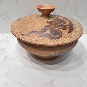 Pode incluir: Uma tigela de cerâmica com tampa, exterior bege salpicado. A tampa apresenta um desenho em espiral castanho escuro e uma pequena pega arredondada. A base da tigela é de uma cor bege semelhante, criando uma estética artesanal. A tigela é adequada para servir ou guardar.