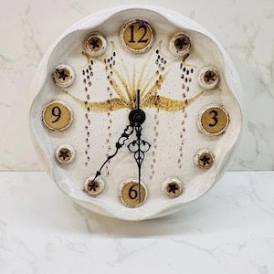 May include: A white ceramic wall clock with a scalloped edge. The clock face features brown numbers and decorative floral accents. The hands are black, and the clock measures approximately 10 inches in diameter.