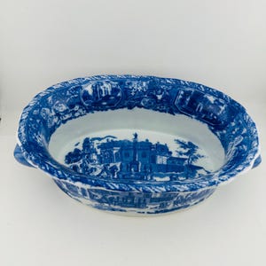 May include: An oval ceramic serving dish featuring a blue and white transferware pattern. The interior and exterior display detailed landscape scenes with buildings and figures. The rim has a scalloped edge and two small side handles.
