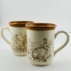 May include: Two ceramic mugs with a cream-colored base and brown rim. Each mug features a brown illustration of a rabbit in a natural setting. The mugs have a curved handle and are suitable for hot beverages.