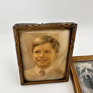 May include: A rectangular antique-style picture frame with a dark metallic finish and cross-hatched corner details, containing a sepia-toned portrait of a young child wearing a collared shirt and tie, positioned next to a second ornate gold-toned frame.