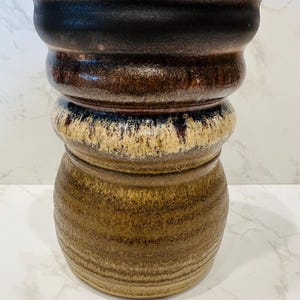 May include: A handcrafted ceramic vase with a unique, layered design. The vase features a dark brown rim, transitioning to lighter brown and beige tones, with a textured, earthy appearance. The vase is approximately 10 inches tall.