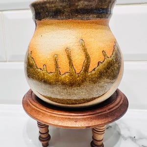 Pode incluir: Um vaso de cerâmica artesanal com corpo arredondado e borda marrom escuro. O vaso apresenta uma paleta de cores quentes de laranja, marrom e bege, e é exibido em um pequeno suporte redondo de madeira.