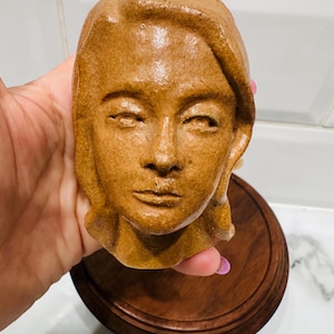May include: A brown ceramic sculpture of a human face with closed eyes and detailed facial features. The sculpture is mounted on a round, dark wooden base. The face has a serene expression and is a warm, earthy tone.