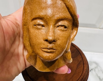 May include: A brown ceramic sculpture of a human face with closed eyes and detailed facial features. The sculpture is mounted on a round, dark wooden base. The face has a serene expression and is a warm, earthy tone.
