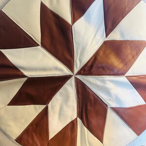 May include: A circular leather cushion featuring a geometric star pattern composed of alternating brown and off-white triangular panels. The pieces are joined with visible stitching, creating a symmetrical design that radiates from the center.