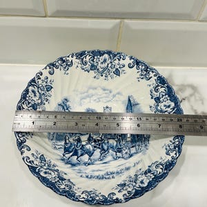 May include: A round white ceramic bowl with a blue transferware pattern featuring a horse-drawn carriage and floral borders. A metal ruler is placed across the center, showing a diameter of approximately 9 inches across the scalloped rim.