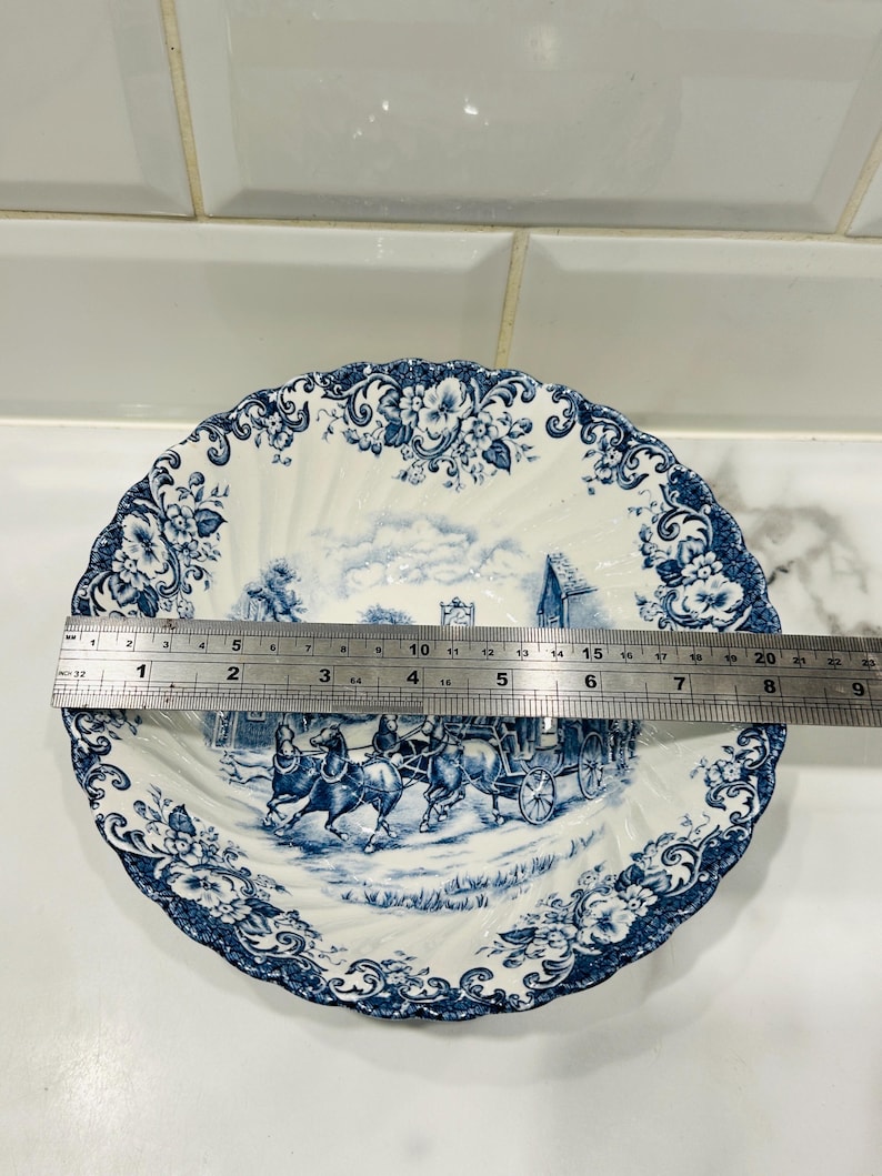 May include: A round white ceramic bowl with a blue transferware pattern featuring a horse-drawn carriage and floral borders. A metal ruler is placed across the center, showing a diameter of approximately 9 inches across the scalloped rim.