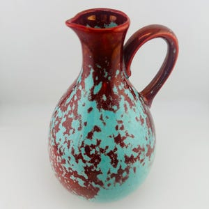 Peut inclure: Un pichet en céramique doté d'un corps bulbeux et d'une anse incurvée. La surface est ornée d'un motif tacheté, semblable à une éponge, composé d'un vernis rouge métallique et turquoise vif, créant un aspect texturé sur tout l'extérieur.