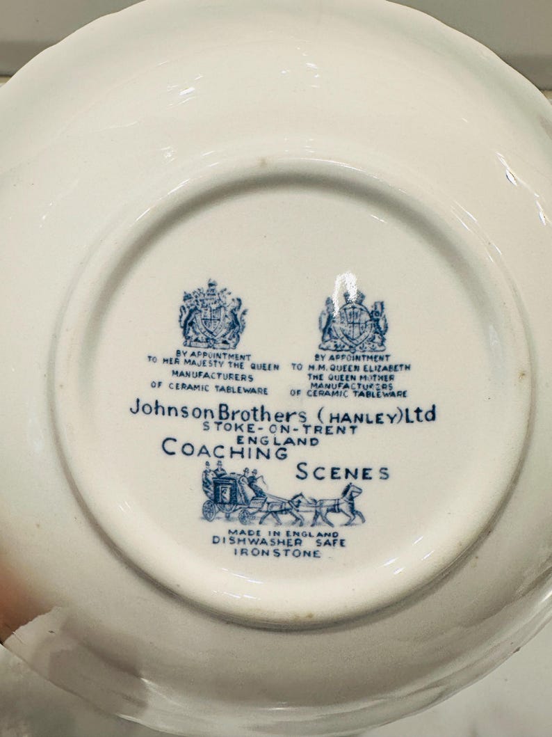 May include: The underside of a white ceramic plate featuring a blue printed maker's mark for Johnson Brothers (Hanley) Ltd, Stoke-on-Trent, England. The text includes Coaching Scenes, Made in England, Dishwasher Safe, and Ironstone.