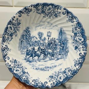 May include: A white ceramic bowl with a scalloped edge and a blue transferware pattern. The center features a detailed scene of a horse-drawn carriage in a village, while the rim is decorated with a dense floral motif of blossoms and leaves.