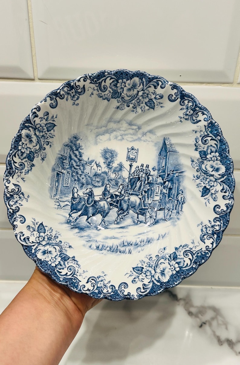 May include: A white ceramic bowl with a scalloped edge and a blue transferware pattern. The center features a detailed scene of a horse-drawn carriage in a village, while the rim is decorated with a dense floral motif of blossoms and leaves.