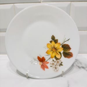 May include: White ceramic plate with a floral design featuring yellow, orange, and white flowers with green and brown leaves. The plate is round and has a simple, elegant design, suitable for serving food.