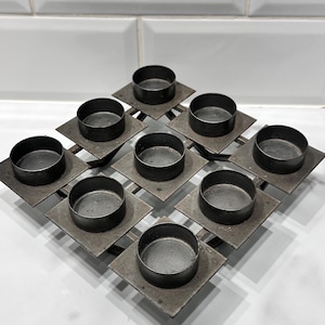 May include: A metal candle holder with nine circular wells. Each well is set on a square base, arranged in a diamond pattern. The metal has a dark, aged finish, suggesting a vintage or industrial style. The candle holder is designed to hold small candles.