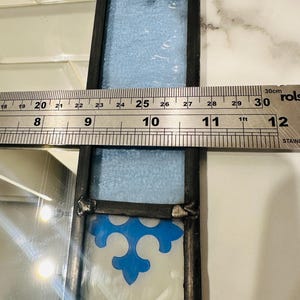 K&ouml;nnte beinhalten: Ein Buntglasfenster mit schwarzem Metallrahmen, bestehend aus einem hellblauen, strukturierten Glasteil und einem wei&szlig;en Glaselement mit blauem Fleur-de-Lis-Muster. Ein Metalllineal zeigt eine Breite von etwa 9 cm an.