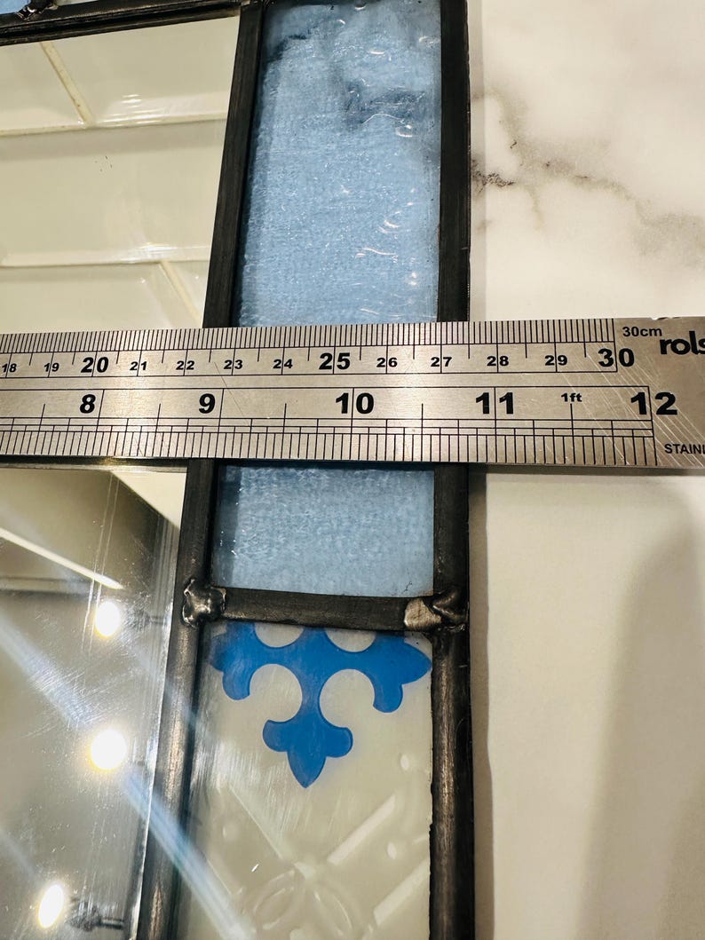 K&ouml;nnte beinhalten: Ein Buntglasfenster mit schwarzem Metallrahmen, bestehend aus einem hellblauen, strukturierten Glasteil und einem wei&szlig;en Glaselement mit blauem Fleur-de-Lis-Muster. Ein Metalllineal zeigt eine Breite von etwa 9 cm an.