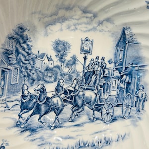 May include: A white ceramic bowl featuring a blue transferware pattern depicting a horse-drawn stagecoach traveling past a village building with a sign, surrounded by floral borders and a scalloped edge design on the rim of the dish.